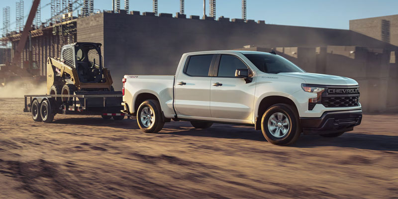 A white Chevrolet Silverado towing a trailer with a skid steer loader through a construction site.